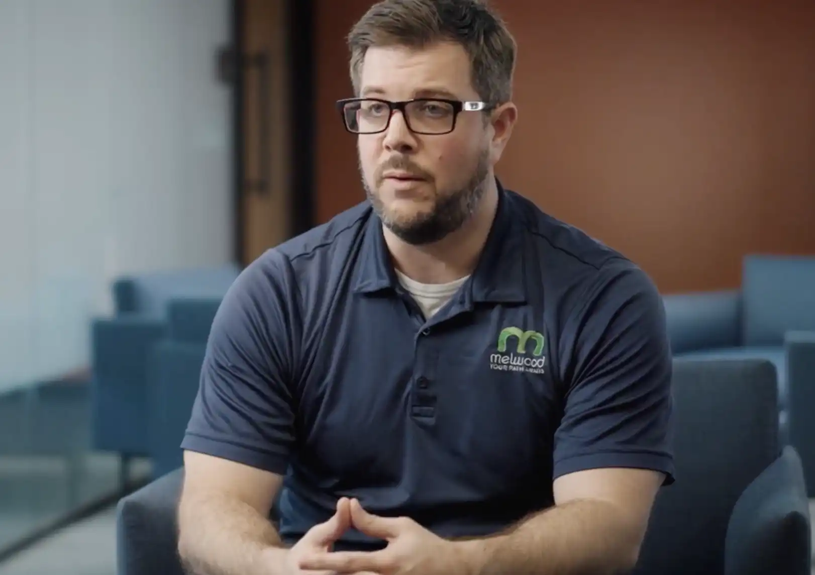 A man wearing glasses and a navy Melwood polo shirt sits in a chair during what appears to be an interview or testimonial video.